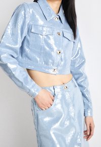 Kort denimjacka i ljusblått med paljetter, guldknappar och två framfickor, ihopparad med en matchande kjol med liknande detaljer.