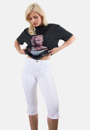 Femme aux cheveux blonds soulevant un T-shirt noir avec l'impression du visage d'une femme souriante, portant un pantalon capri blanc ajusté sur un fond uni.