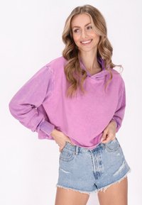 Felpa viola, oversize, a maniche lunghe con collo a cowl, abbinata a pantaloni corti in denim chiaro e strappato. Tessuto morbido, stile casual.