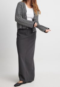 Cardigan rayé noir et blanc, à l'avant ouvert, associé à une longue jupe gris foncé et des ballerines noires, avec des manches longues dotées d'ouvertures pour les pouces.