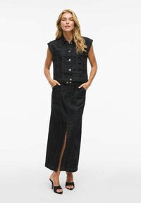 Svart denim utan ärmar jacka i kombination med en lång kjol med en slits framtill. Båda plaggen har en strukturerad design och synlig sömnad.