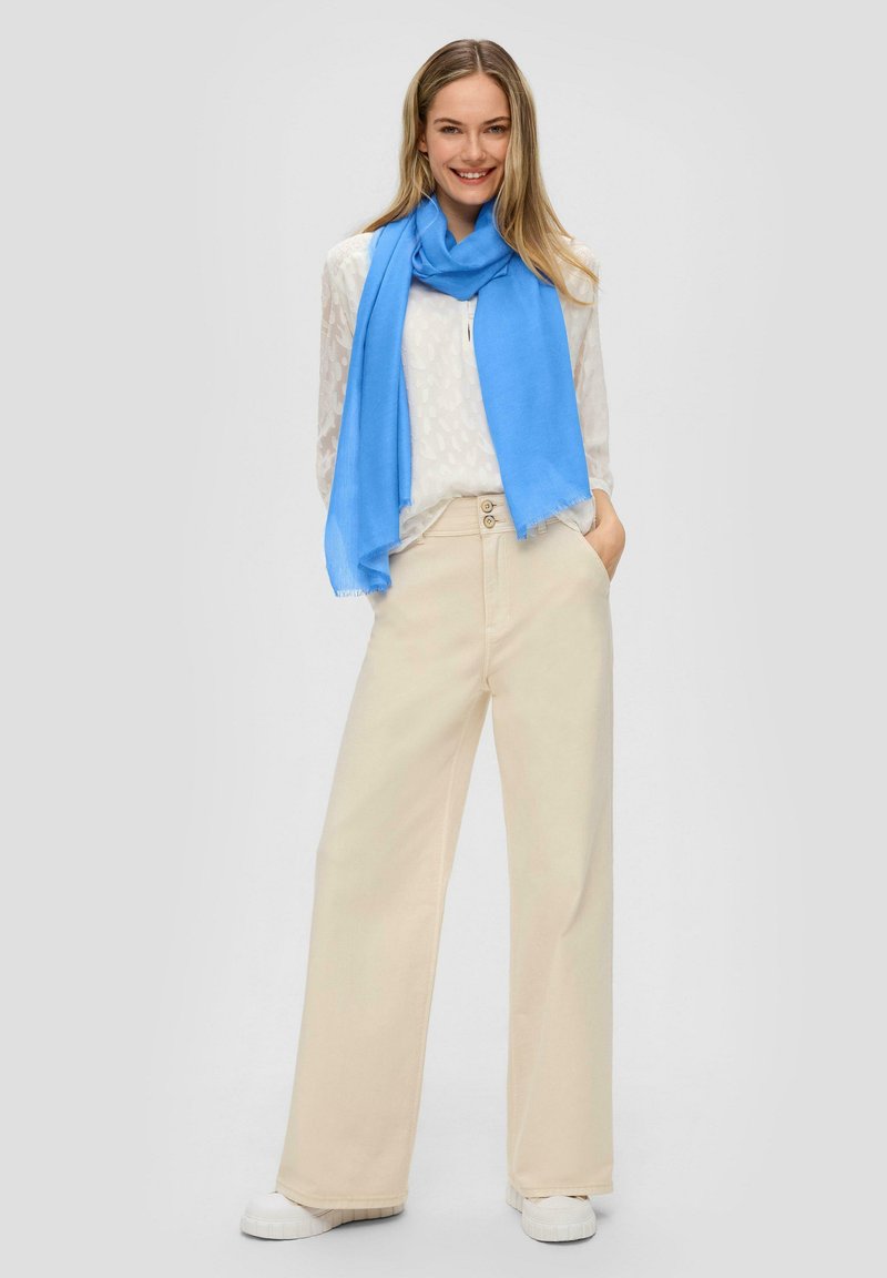 Écharpe bleu clair, blouse blanche à motifs, pantalon beige à jambes larges, baskets blanches. Texture lisse, coupe décontractée. Détail de taille à deux boutons.