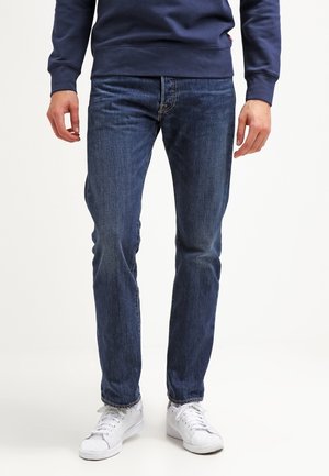 Ein Mann trägt dunkelblaue Jeans, ein marineblaues Sweatshirt und weiße Turnschuhe und steht mit entspannten Armen seitlich am Körper.