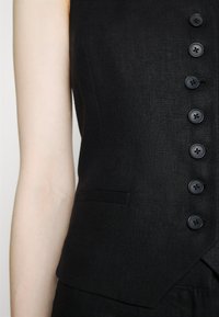 Gilet noir sur mesure avec sept boutons et une petite poche avant porté par une personne à la peau pâle visible sur le bras.