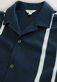 Marineblaue, strukturierte Hemdjacke mit ausgestelltem Kragen und zwei vertikalen weißen Streifen auf der rechten Seite.