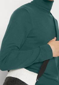 Türkiser, gerippter Rollkragenpullover mit langen Ärmeln, getragen während eine weiße und schwarze, strukturierte Handtasche mit Schulterriemen gehalten wird.