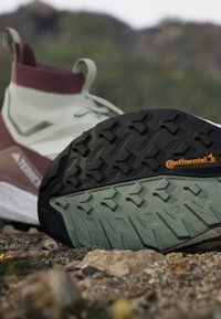 Polkujuoksukengät, joissa on teksturoitu musta kumipohja, vihreä ja harmaa välikerros sekä valkoinen ja burgundinpunainen päällinen, joka sisältää sukkamaisen muotoilun.