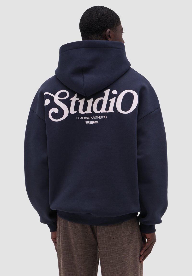 Man draagt een marineblauwe hoodie met grote witte tekst "Studio" en kleinere tekst "Crafting Aesthetics WRSTBHVR" op de achterkant, gecombineerd met een bruine broek.