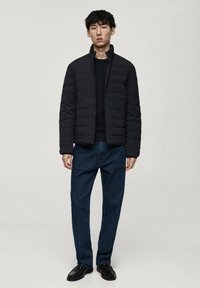 Veste noire matelassée avec un col montant, portée sur un pull sombre. Le modèle porte un jean noir et des chaussures noires. Design simple et moderne.