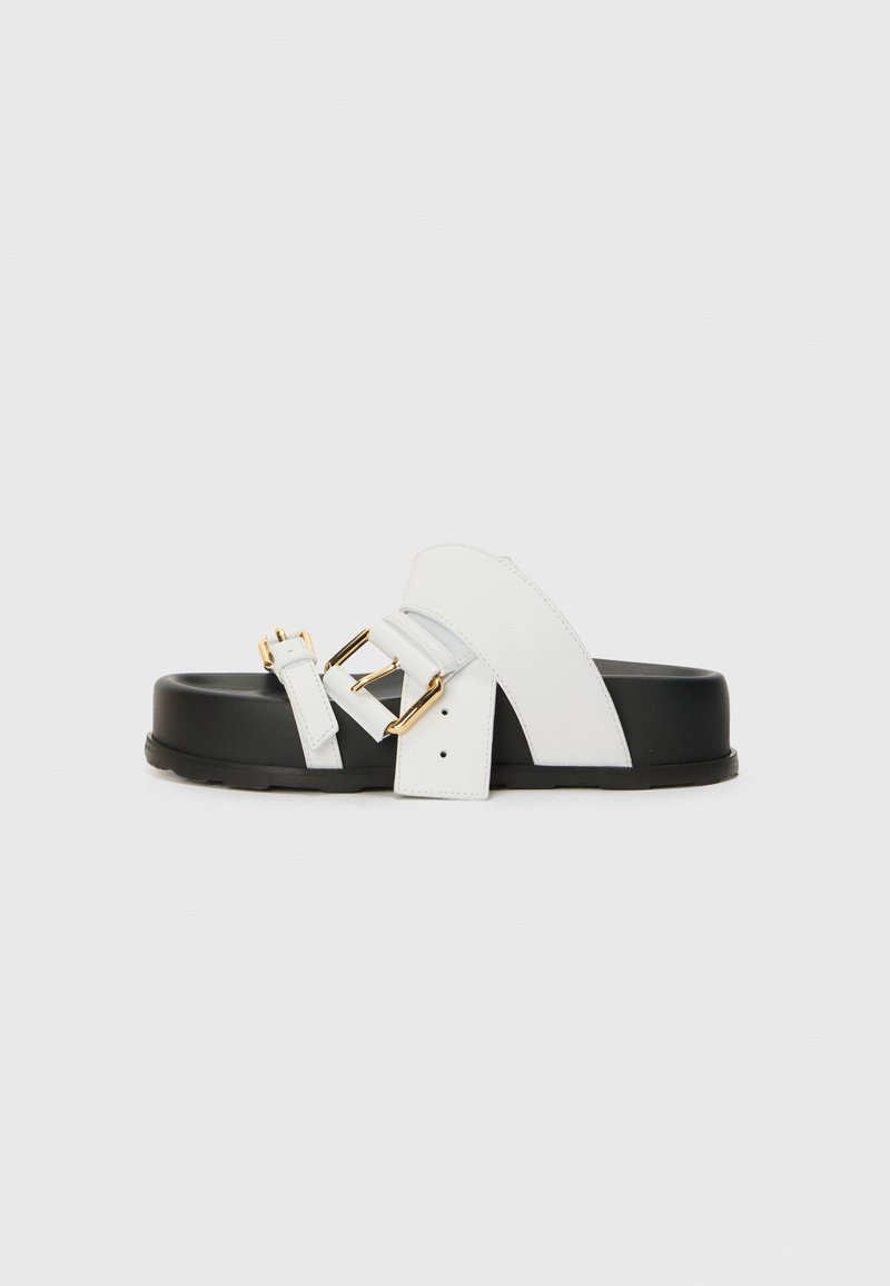 White leather sandals with a wide strap and gold buckles, resting on a black platform sole. Features smooth and matte textures.