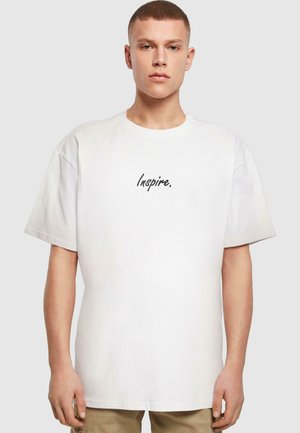 Hvid kortærmet t-shirt med centreret sort kursiv tekst, der siger "Inspire." Fremstillet af blødt bomuld med en løs pasform.