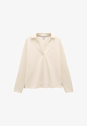 Krémová košeľa s dlhým rukávom, otvoreným golierom a jemne textúrovanou látkou, s malými rozparkom na bočných stranách lemu a slabým logom na ľavej hrudi.