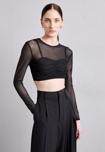 Svart langermet topp laget av gjennomsiktig stoff med rynket, kort bodice og tynne stropper, kombinert med høyt liv svarte bukser.