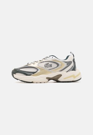 Zapatilla deportiva con paneles de malla y sintéticos en blanco, beige y verde oscuro, suela gruesa y un pequeño logo de cocodrilo en el lateral.