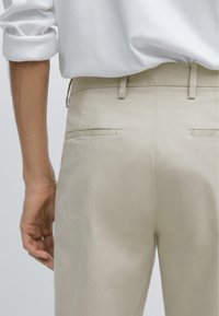 Pantaloni beige realizzati in tessuto liscio, caratterizzati da una vita aderente, passanti per la cintura e due tasche posteriori. La cintura è piatta con cuciture sottili.