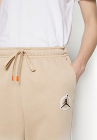 Beiga sweatpants med elastisk midja, snörning med orange spetsar och en vit cirkulär logotyp med en svart silhuett av en hoppande figur.