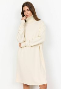 Vestido de punto color crema con un alto cuello acanalado, mangas largas y un corte oversize, que cae justo por encima de las rodillas.