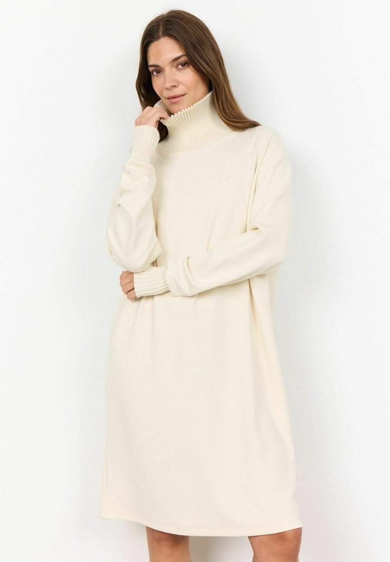 Vestido de punto color crema con un alto cuello acanalado, mangas largas y un corte oversize, que cae justo por encima de las rodillas.