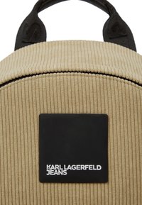 Rucsac din corduroy bej, cu un vârf rotunjit, având o etichetă dreptunghiulară neagră cu inscripția "KARL LAGERFELD JEANS" embosată în litere albe.