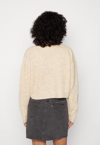 Pull en tricot beige avec une coupe courte, associé à une jupe en denim gris foncé avec poches arrière et une étiquette logo rouge.