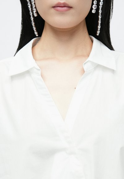 Camisa de colarinho branco com uma gola dobrada e textura suave; adornada com brincos longos de cristal brilhante.
