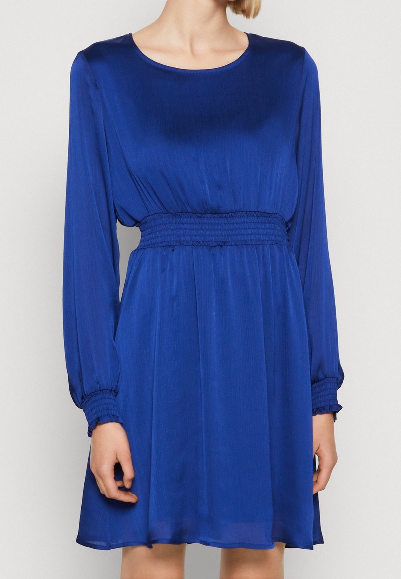 Robe bleu roi à manches longues avec un col rond et une taille froncée. Tissu fluide avec un léger éclat, se terminant au-dessus du genou.