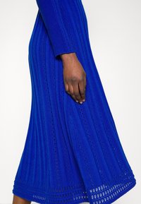 Robe en tricot bleu vibrant avec une texture côtelée verticale et des détails en dentelle. Manches longues avec une finition évasée.
