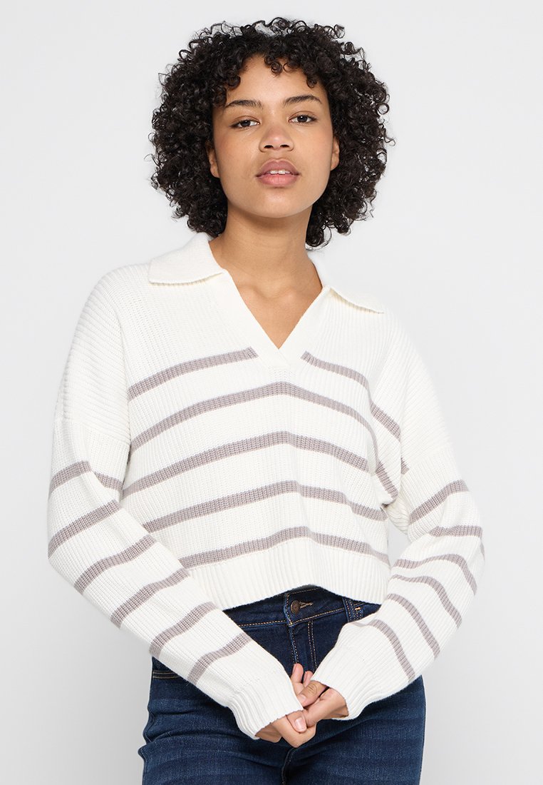 Kobieta z kręconymi włosami ubrana w biały sweter z kołnierzykiem w szare poziome paski oraz ciemnoniebieskie dżinsy, stojąca na tle białym.