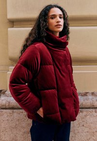 Red velvet padded jacket with a high collar, featuring a soft texture and a relaxed fit. The design includes horizontal quilting and side pockets.