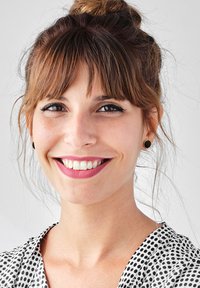 Donna con capelli castani raccolti a chignon, che indossa una blusa bianca con pois neri, orecchini a bottone neri e un rossetto di colore rosa chiaro.