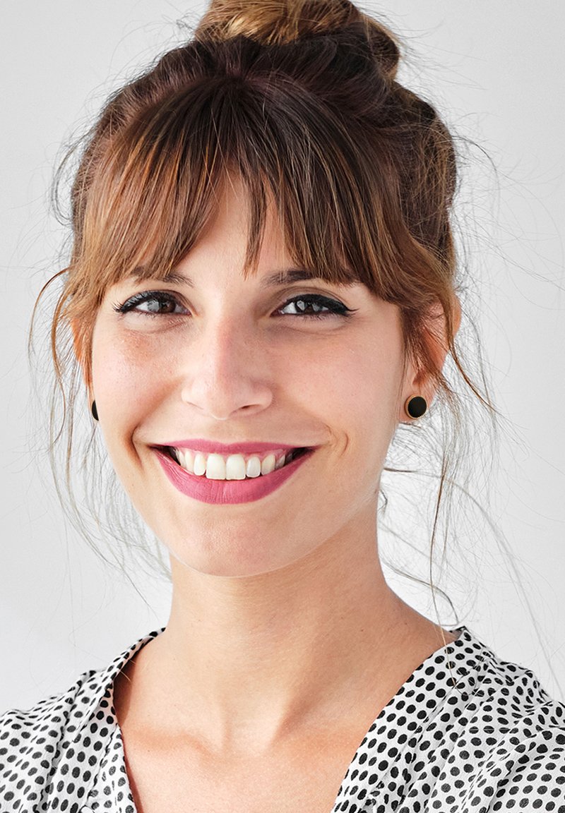 Donna con capelli castani raccolti a chignon, che indossa una blusa bianca con pois neri, orecchini a bottone neri e un rossetto di colore rosa chiaro.