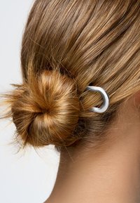 Vaaleat hiukset on muotoiltu alas buniksi, joka on kiinnitetty hopeisella D-muotoisella hiussolmulla neutraalia taustaa vasten.