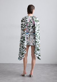 Robe courte à motifs floraux avec une cape fluide verte et blanche, ornée d'accents roses et orange, accompagnée de sandales à talons blancs.