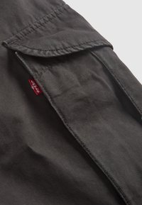 Zwarte stoffen shirt met een klepzak, zichtbare stiksels en een klein rood merkembleem nabij de zakopening. Zachte, gladde textuur.