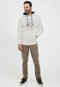 Hvid hættetrøje med sort hætte og trykt logo, parret med beige bukser og sorte sneakers. Har en lomme foran og snore.
