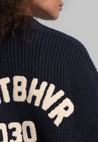 Marineblau gestrickter Pullover mit creamfarbenem besticktem Text, gerippter Textur und lockerer Passform. Zu den Details gehören übergroße Buchstaben und ein weit gefasster Kragen.