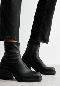 Botas de tobillo de cuero negro con textura suave, caña corta, tacón en bloque inclinado y detalle de cremallera lateral. Llevadas con jeans oscuros.