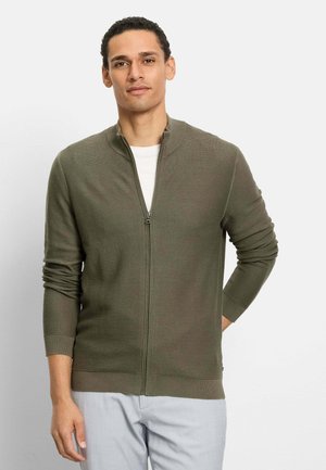 Mann trägt olivgrünen Reißverschluss-Pullover über weißem Hemd und hellgrauer Hose, steht mit einer Hand hinter dem Rücken.