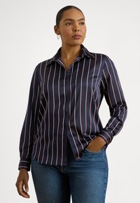 Žena v tmavomodrej blúzke s bielymi vertikálnymi pruhmi a modrých džínsach, pózujúca s jednou rukou v vrecku na bielom pozadí.