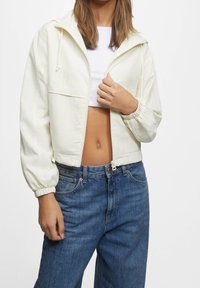 Veste blanche coupée avec fermeture éclair sur le devant et capuche, munie de poignets élastiques et d'une coupe décontractée, associée à un jean en denim bleu taille haute.