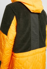 Quiltad jacka med orange och mörkgröna sektioner, som har ett strukturerat fleece-material och en slät svart detalj på baksidan.