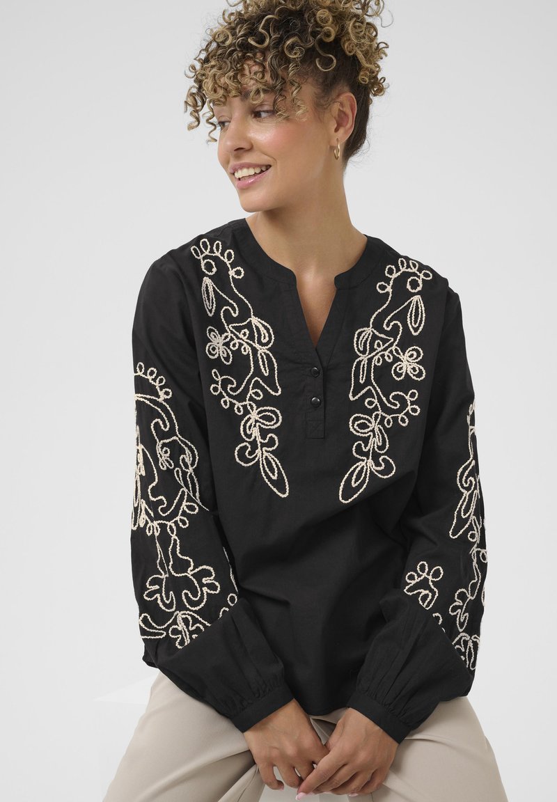 Zwarte blouse met lange mouwen, voorzien van ingewikkelde beige bloemenborduring op de mouwen en de voorkant, met knoopsluiting bij de nek.