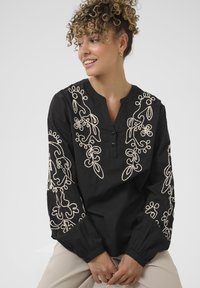 Sort langærmet bluse med detaljeret hvid blomsterbroderi langs ærmerne og halsudskæringen, med knaplukning foran og plisserede manchetter.