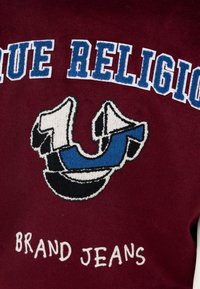 Bordowy materiał z haftowanym logo. Zawiera duży niebiesko-czarny wzór z białymi akcentami. Napis brzmi "TRUE RELIGION BRAND JEANS."