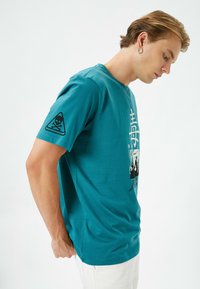 Teal bomulds T-shirt med et grafisk design med japansk tekst og et kranium emblem på ærmet. Korte ærmer og rund hals.