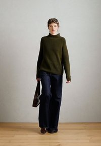 Maglione lavorato a maglia verde scuro con collo alto, abbinato a jeans a gamba larga blu scuro. Il modello tiene in mano una borsa marrone, in piedi su un pavimento di legno.