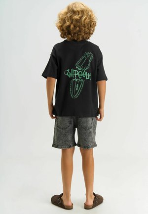 Sort T-shirt med korte ærmer og et grønt grafisk motiv på ryggen, kombineret med grå denimshorts og brune slip-on sko.