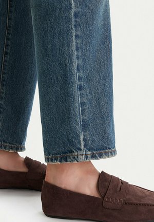 Füße in braunen Wildlederloafern, kombiniert mit blauen Denim-Jeans, die oberhalb der Knöchel gekürzt sind, vor schlichtem Hintergrund.