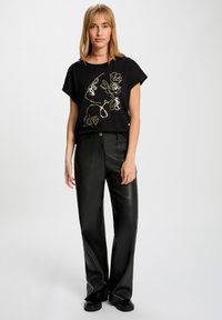 Camiseta negra con un diseño gráfico dorado que presenta contornos florales y texto, combinada con pantalones de imitación cuero negros de tiro alto.