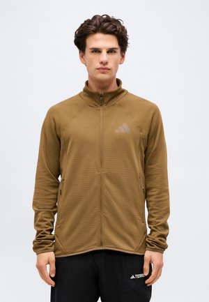 Młody mężczyzna ubrany w brązową kurtkę na zamek z logo Adidas oraz czarne spodnie, stoi na tle jednolitego białego tła.